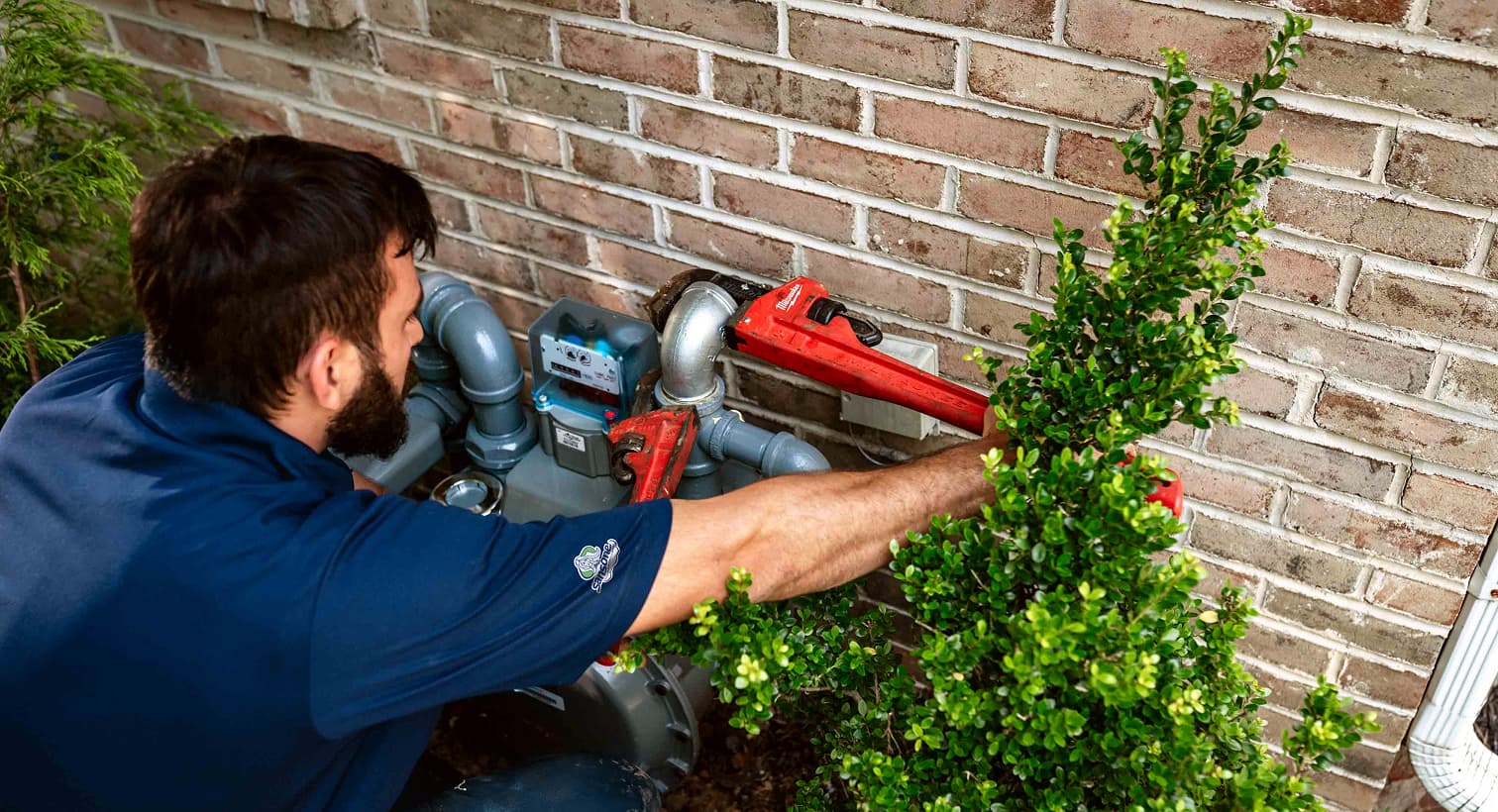 A professional Sansone technician using a red pipe wrench to service outdoor residential gas line plumbing and pipe connections against a brick wall.