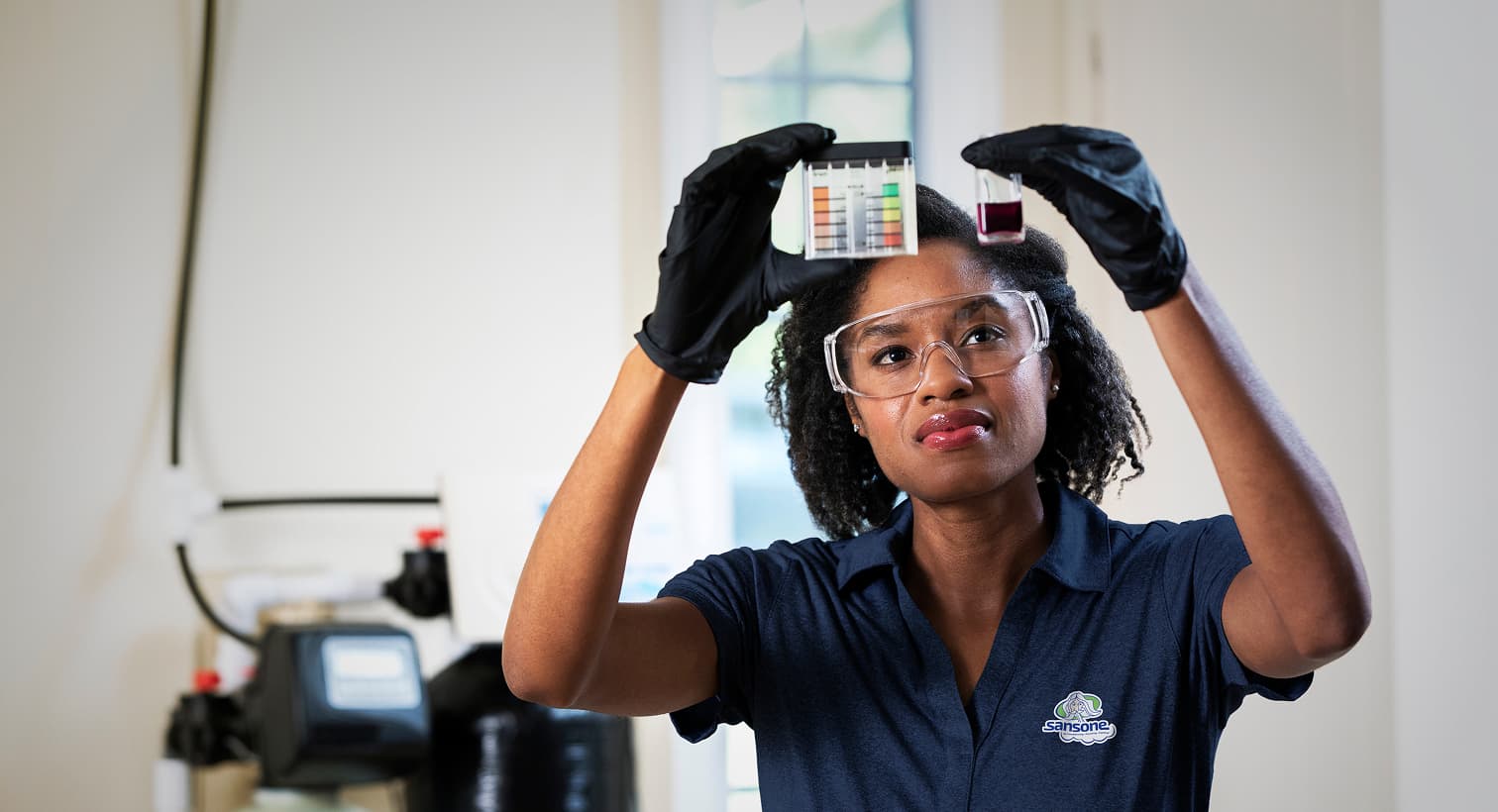 A Sansone technician in a blue uniform and safety gear compares a water sample to a testing chart in front of a water softener system.