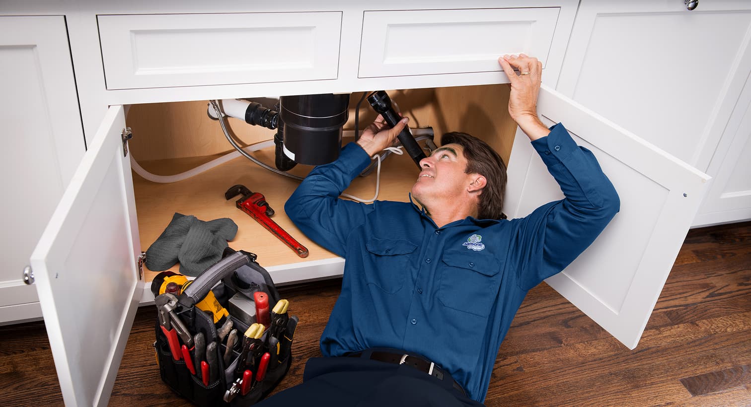 A professional Sansone plumber using a flashlight to inspect pipes under a kitchen sink for residential plumbing leak detection services.